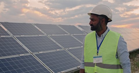 Mixed ethnicity male engineer in uniform walking and looking at solar power plant. Bearded man in hard helmet examining object. Concept of solar station development and green energy Arkistovideo
