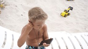 Blonde child sitting on the beach on a Sunny summer day listening to music with a smartphone and headphones. Dependence on gadgets.