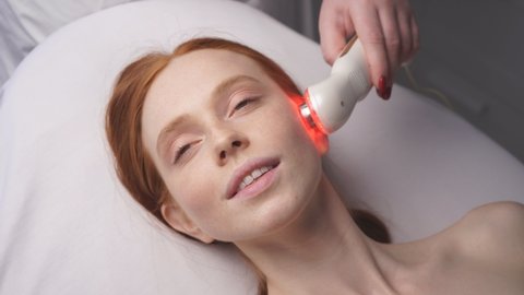 attractive woman in a cosmetology clinic receives a phonophoresis procedure for the skin of the face. The cosmetologist moves a manipulator with a blue light on the face.