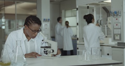 Black Female Laboratory Scientist Conducts Experiment in Science lab looking at microscope in busy lab at night