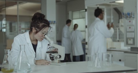 Asian Female Laboratory Scientist Conducts Experiment in Science lab looking at microscope in busy lab, diversity