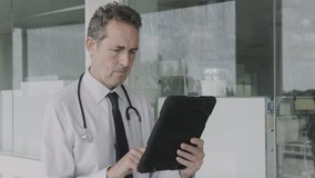 Mature Male Doctor standing in modern hospital corridor working on digital tablet and looking into hospital ward - Powered by Shutterstock - Get 15% off with code: PIKWIZARD15