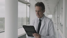 Mature Male Doctor standing in modern hospital corridor working on digital tablet and looking into hospital ward - Powered by Shutterstock - Get 15% off with code: PIKWIZARD15