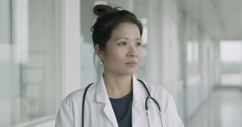 Portrait of Young Adult Female Chinese Doctor standing in modern hospital corridor looking at camera pensive, wearing lab coat and stethoscope