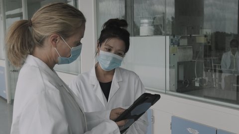 Female doctors meeting in hospital corridor discussing test results on digital tablet computer technology, wearing surgical face mask during Coronavirus pandemic