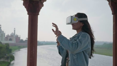 A happy young attractive woman or female in the jacket is standing outside in front of the Taj Mahal and making hand gestures and watching 360 video wearing 3D glasses VR or Virtual reality headset. 