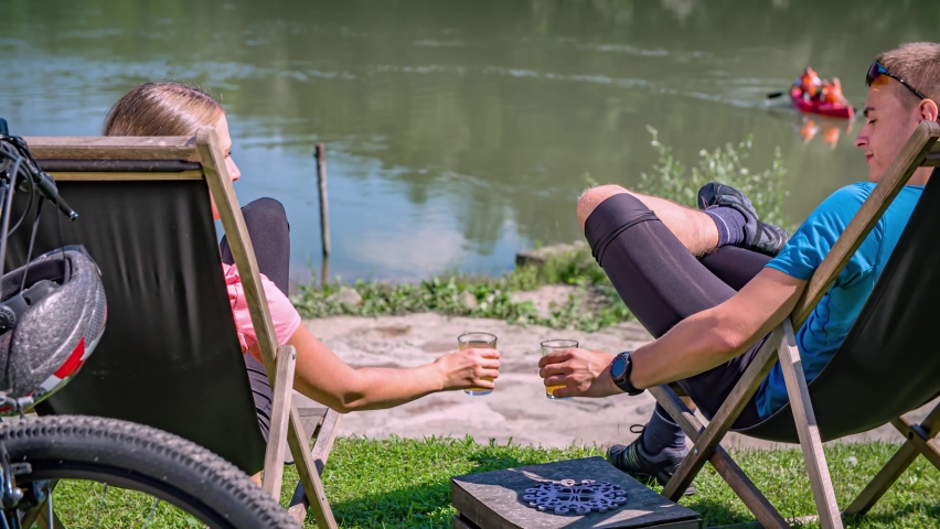Couple enjoy drinks while resting in chairs on riverbank after bike ride.