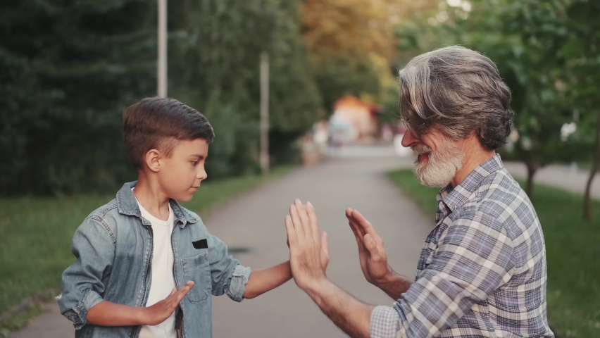 Grandfather in a stylish look and his little grandson are playing with hands in a park. Close view.