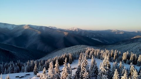 Drone flying above mountain valley in winter. Aerial view of snow covered pine forest hills at sunrise with huts on meadow