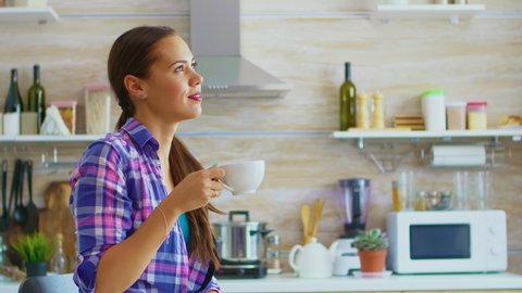 Young lady drinking green tea and smiling at breakfast sitting at the table in the kitchen. Woman enjoying healty herbal hot drink in the morning using teapot and teacup.