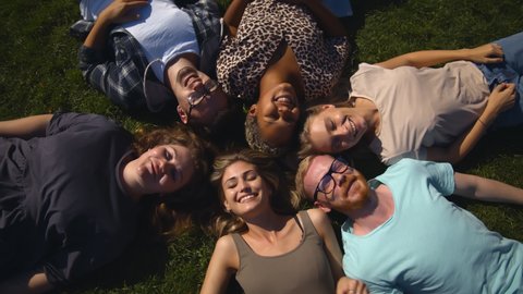 Top view of diverse team of friends relaxing in park lying on grass in circle. Happy smiling students resting or green lawn outdoors in park. Friendship, diversity concept