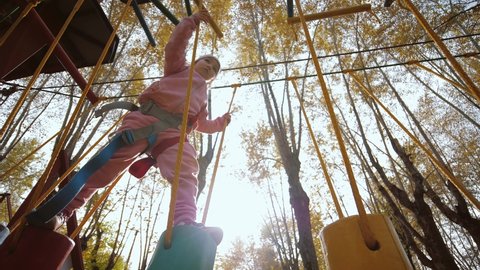 Little girl walking and balancing on swinging wooden stump in rope park. Cute brave child overcome obstacle course, make steps, entertainment in adventure rope town, spending time outdoors