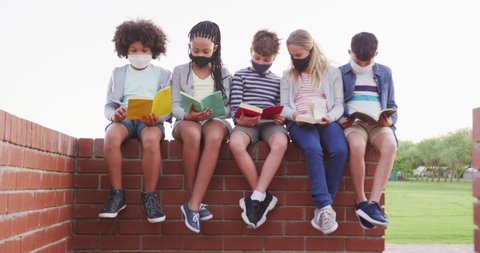 Group of multi ethnic kids wearing face masks reading books, sitting on the wall during a break. Primary education social distancing health safety during Covid19 Coronavirus pandemic in slow motion. Arkivvideo