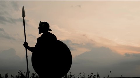 Silhouette of spartan in red cloak and armor posing with shield looking around, beautiful sunset on background. Side motion of incognito roman gladiator holding spear outdoors. Concept of warrior.