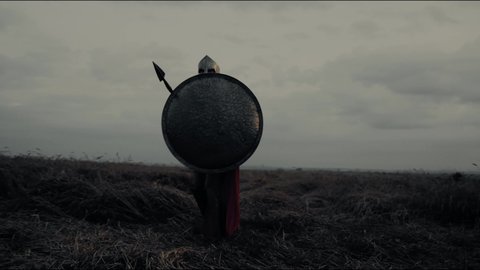 Muscular shirtless spartan in red cloak and armor holding shield. Drone motion of roman gladiator in iron helmet with sword on belt practicing attack pose using spear in field with dry grass, evening.