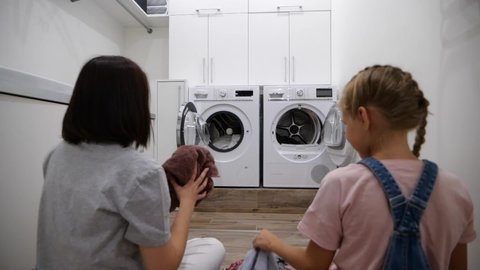 Young woman and her daughter having fun while doing laundry at home throwing clothes into washing machine. Slow motion