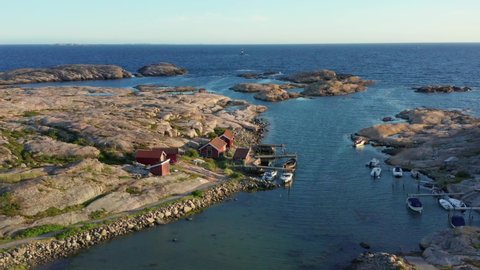 Swedish Fishing houses on west coast at blue ocean. Circling drone flight close over water surface red cottage shed. Small fisher boats at pier. Sunny evening light rocks and stones. Sweden coastline