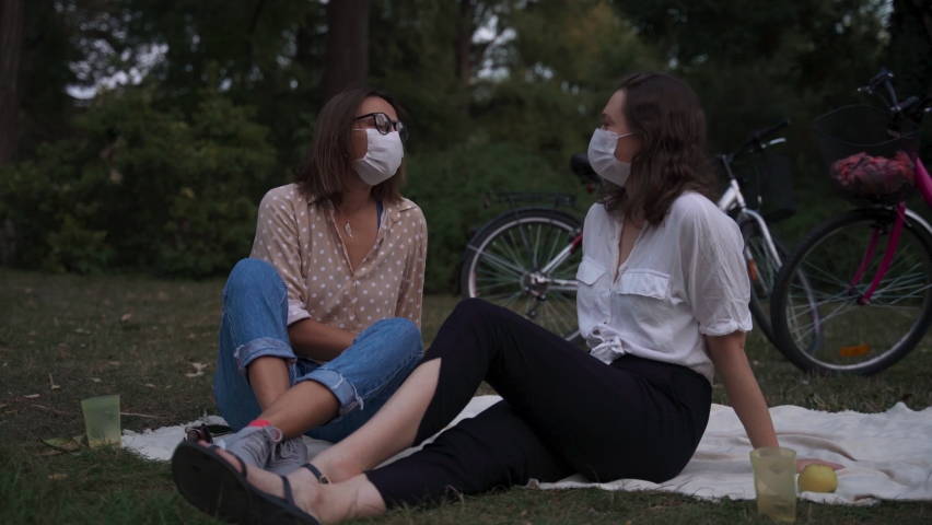 Two young women in protective face masks sitting on a blanket at the park talking and laughing while on a picnic. New normal concept.