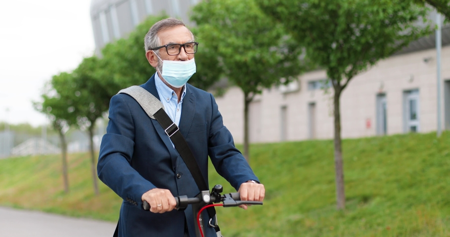 Old gray-haired man in glasses and medical mask standing on electric scooter and riding at street in town. Senior male rushing somewhere and looking at hand watch outside. City vehicle concept.
