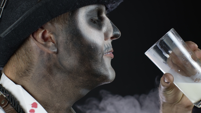Side view of creepy man with skeleton makeup in white shirt with blood. Guy looking at camera, drinks milk from a glass. Voodoo rituals. Halloween thematic party. Black background. 6k downscale