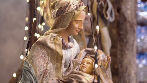 Close-up of Holy Mary and Jesus Christ figurines decorated with illuminating garland. Jesus Nativity scene, mother holding newborn savior child in hands in stable. Holy night and christmas decorations
