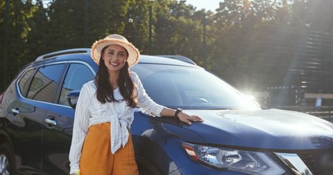 Outdoor portrait of happy young woman standing near her new car and showing keys, sun flare