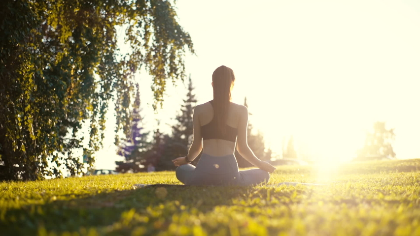 Back view of unrecognizable slender young woman sitting on yoga mat in lotus position and raising hands up outside in city park. Rear view of female practicing yoga outdoors in sunny day. | Shutterstock HD Video #1059824771
