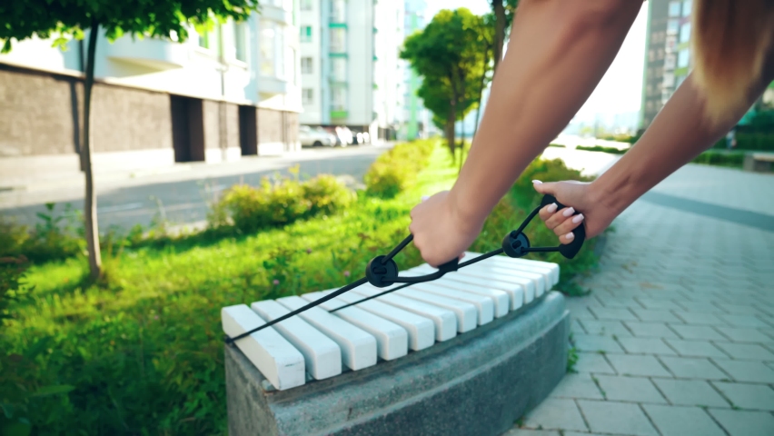 Close up of arms of unrecognizable woman training upper body, practicing slow controlled exercises using fitness elastic rope band. Girl enjoying working with resistance band on bench in city street.