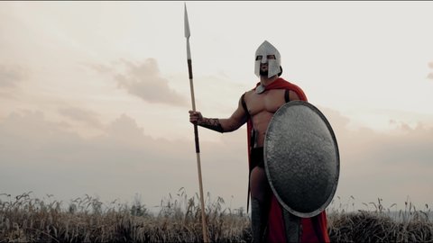 Side motion of roman gladiator with sword on belt holding spear in dry field. Muscular shirtless spartan in red cloak and armor posing with shield looking at camera, beautiful summer sunset.