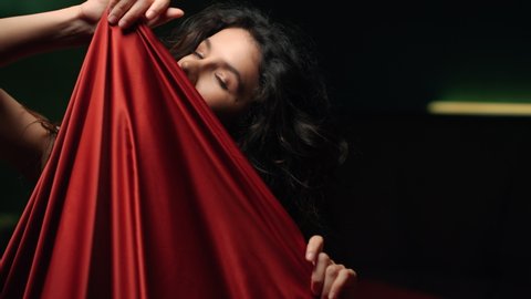 Portrait of sexy woman playing with red silk cloth. Closeup nude girl taking erotic poses with silk sheet indoors. Seductive girl flirting and posing for camera in dark bedroom.