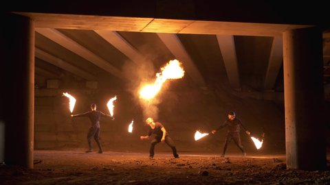 Two fire show artists come out from behind columns with fire chains. The third fire show artist breathes fire. An abandoned place. Impressive performance. Slow motion
