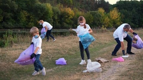 A family of volunteers with children clears the river Bank of garbage. People collect plastic leftovers in garbage bags. Environment, ecology, biosphere protection concept.