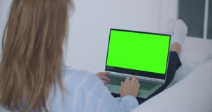 A young woman lying on the couch looks at the screen of the laptop with a green screen and nods her head. Make a video call