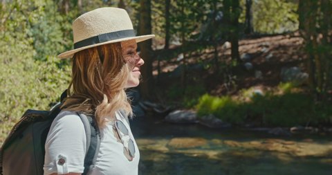 Slow motion beautiful female traveler enjoying summer sunny day. Smiling woman with nature and green forest on background. Traveler with backpack staying at forest creek, smiling and feeling happy, 4K