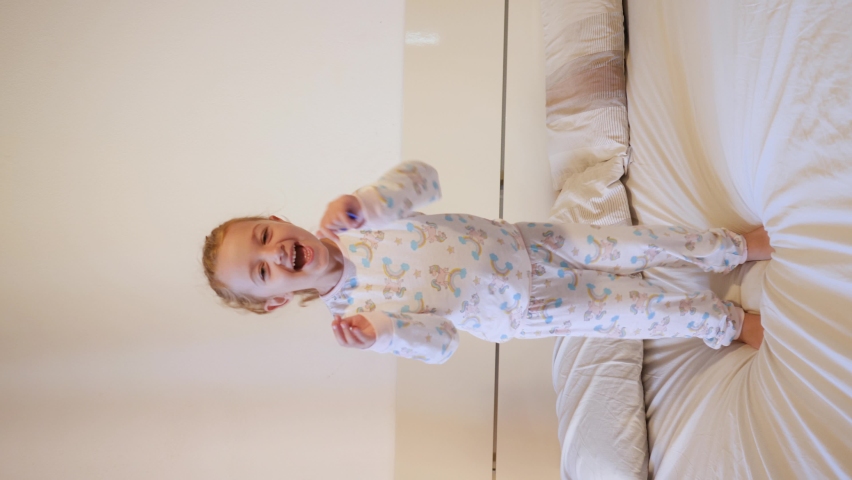 VERTICAL VIDEO - 4 year old Caucasian girl jumps and laughs on the bed in unicorn sleepwear pajamas. Royalty-Free Stock Footage #1059842567