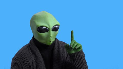 Old retired alien in warm knitted jacket instructively raises index finger in the air and nods his head against blue chromakey background. Themed costume for party