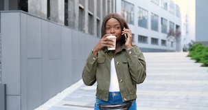 African American joyful young stylish woman talking on cellphone and sipping hot drink, walking in the morning at street. Beautiful happy female speaking on mobile phone and drinking coffee. Outside.