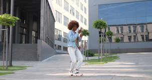 Joyful young African American beautiful curly woman in headphones dancing at city street and listening to music on smartphone. Pretty happy female enjoying song and moving like cool dancer.