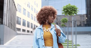 Portrait shot of African American young hipster pretty woman in jeans jacket standing at city street, turning face and smiling joyfully to camera. Beautiful curly happy cheerful female outdoors.
