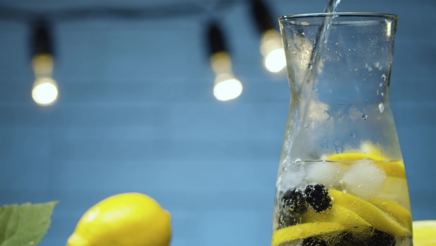 Extreme close up dewberry lemonade. Pouring soda water into a jug with dewberry, sliced lemon and ice. Blurry lamps on blue background. Slide or move camera, dolly shot