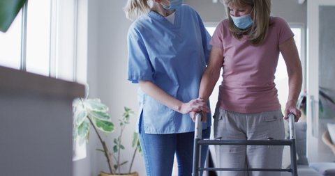 Senior Caucasian woman at home visited by Caucasian female nurse,
walking using a walker, nurse wearing face mask. Medical care
at home during Covid 19 Coronavirus quarantine, slow motion