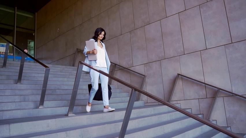 Young confident brunette business woman in blue suit walking down stairs with laptop, documents and big black bag. Busy beautiful girl leaving office building after work.