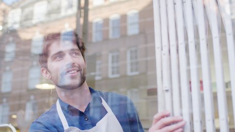 Man running small business pushing back security grill from window and opening shop viewed from outside - shot in slow motion