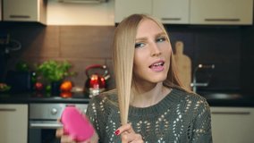 Young vlogger combing hair for camera. Pretty young woman brushing blond hair with comb while shooting video for beauty vlog in cozy kitchen.