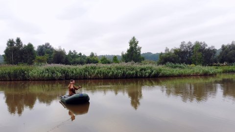 young man on an inflatable boat sits and throws a spinning or fishing rod into the water in the middle a lake or river. background of reeds and forest. Hipster guy fisherman active recreation, camping