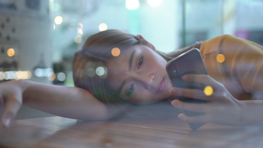 4K Loneliness young beautiful woman sitting by the window lying on the table and using smartphone with internet in cafe at night. Sad teenage girl looking out of the window to city street night lights Royalty-Free Stock Footage #1059855659