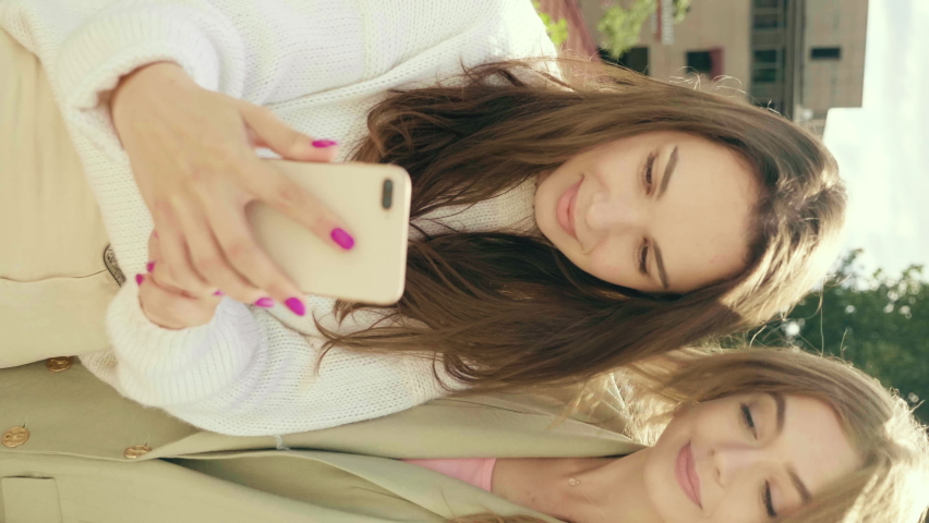 Vertical video.Two young beautiful smiling hipster girls.Sexy carefree women posing in the street in hats. Models having fun.They looking at cellphone screen laughing enjoying using mobile apps
