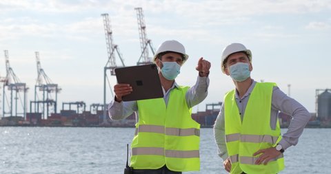 Port workers work with a tablet and point to the object in front of them, standing against the backdrop of marine cranes. Engineers improve seaport logistics.