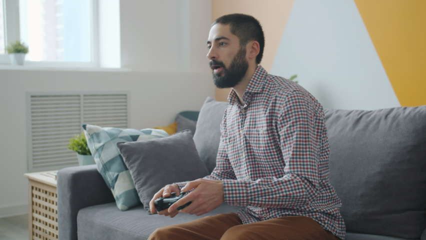 Carefree Middle Eastern guy is enjoying video game at home using joystick relaxing on couch at home alone. Youth culture and entertainment concept. | Shutterstock HD Video #1059857696