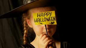 Little pretty girl witch in black wizard hat holds greeting Happy Halloween card says Boo and laughs. Child dressed in ghost party costume scarily staring at camera 4K festive autumn holiday footage.